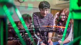 Close up image of a group of students working on a drone. A green light can be seen reflecting off of the glass in front of the students..